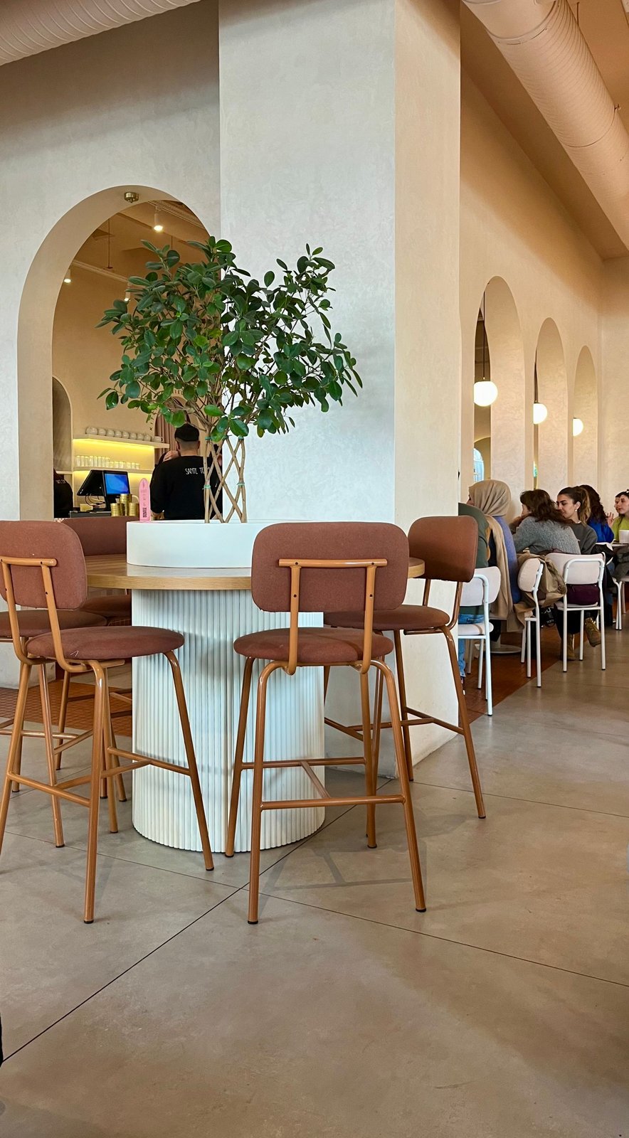 A warm cream cafe with arched openings, a fluted counter, wooden stools and indoor trees.