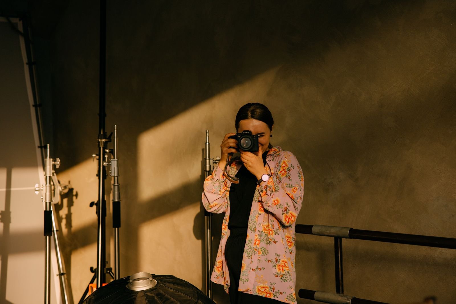 A photographer holding a camera in a warmly lit studio with dramatic diagonal sunlight.