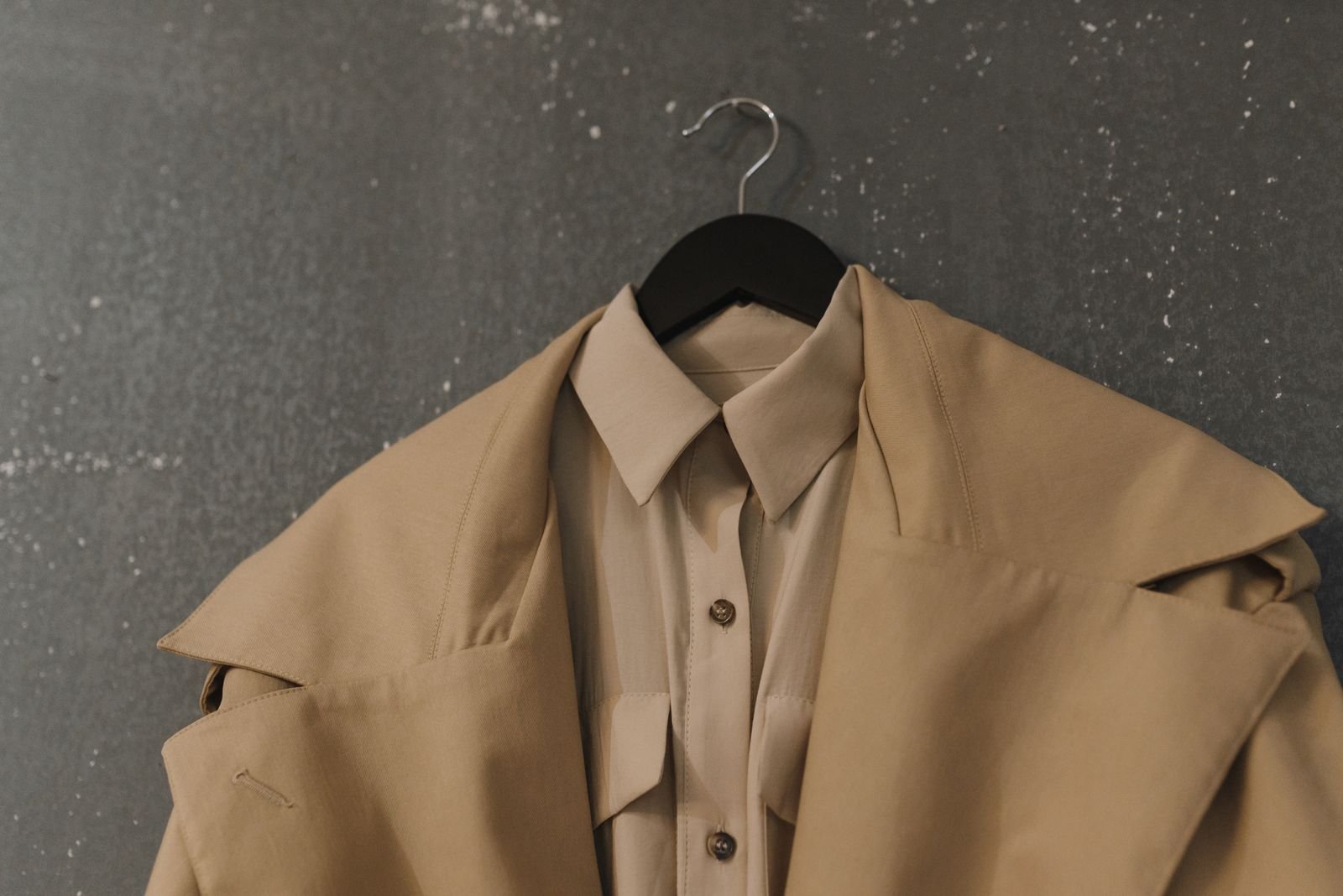 A beige collared shirt and trench coat hanging together on a black hanger against a textured gray wall.