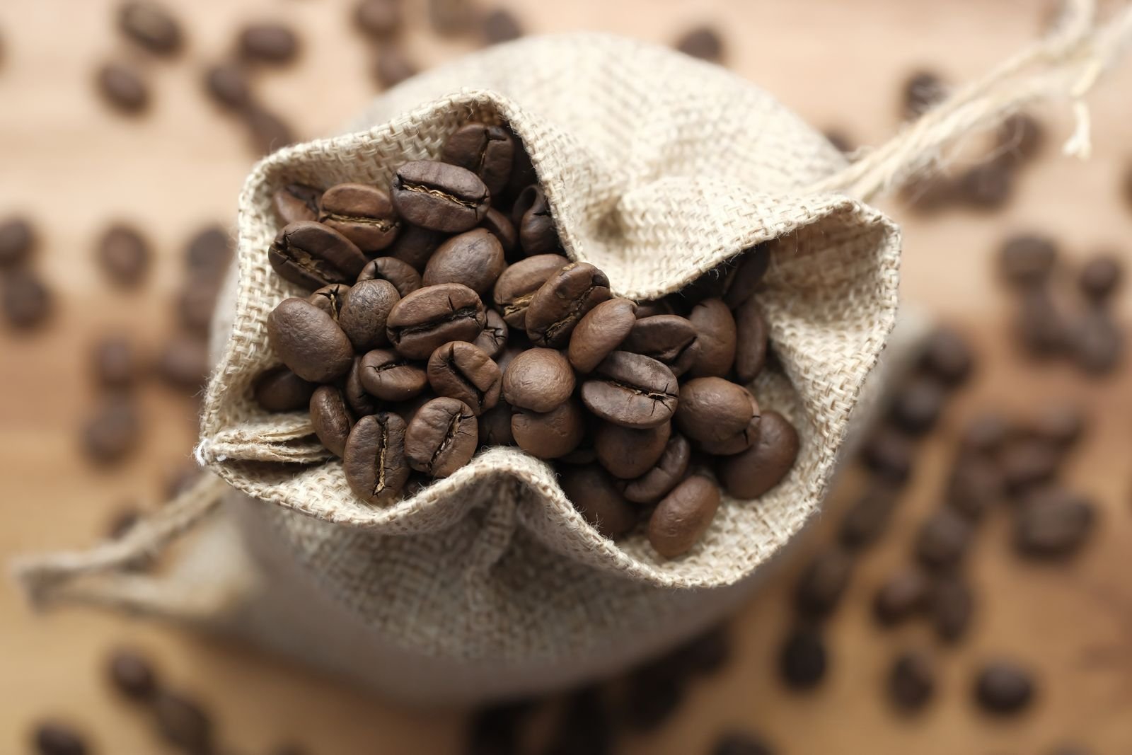 Roasted coffee beans spilling from a small open burlap sack onto a warm surface.