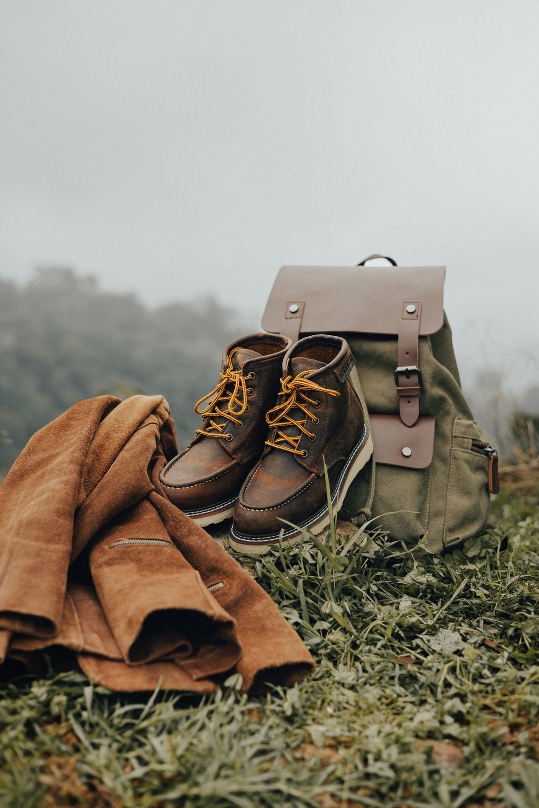 A green canvas backpack, brown leather boots and a folded brown jacket on grass in a misty landscape.