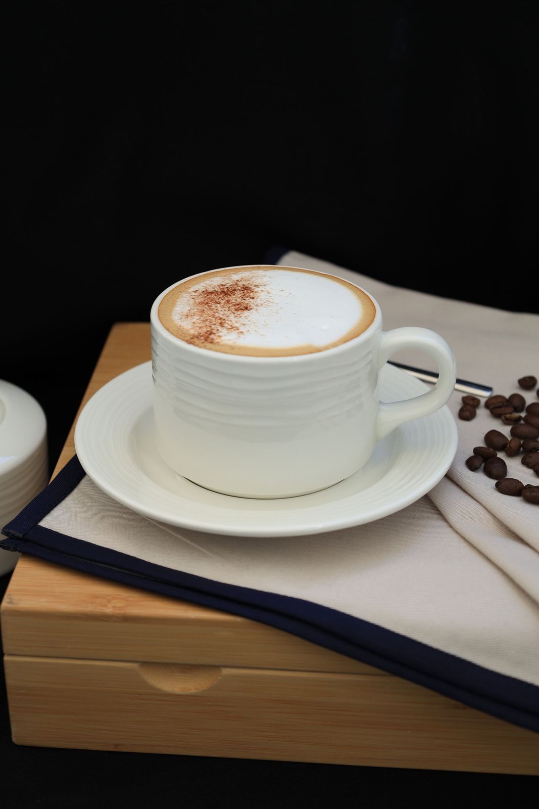 A white cup and saucer holding a foamy coffee, styled with beans and a napkin on a wooden box.