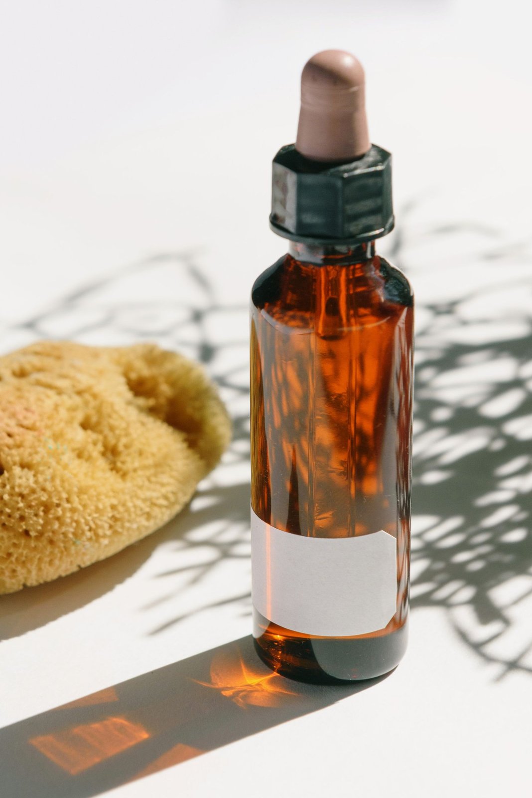 An amber glass dropper bottle with a blank white label beside a natural sponge in direct sunlight.
