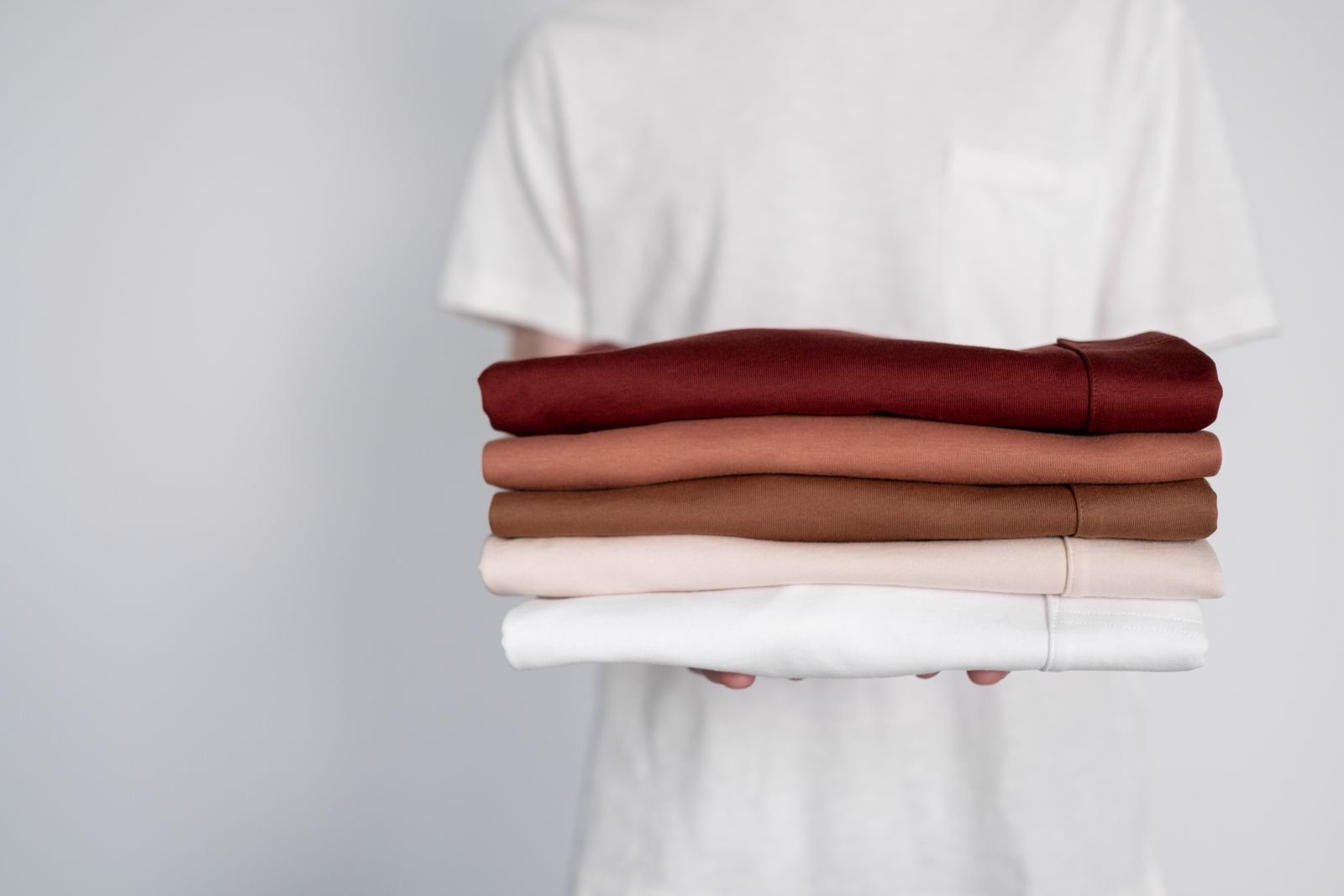 A person in a white shirt holding a neat stack of folded garments in burgundy, rose, beige and white.