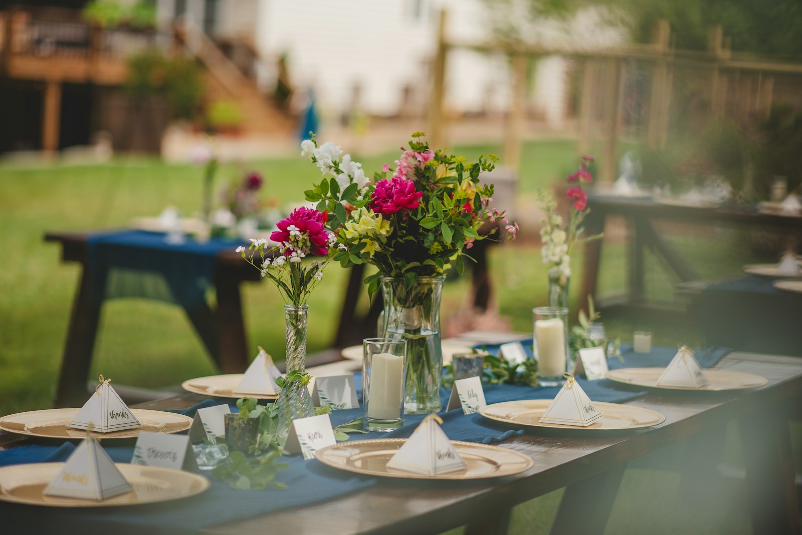 Styled wedding reception table with pink and white floral centerpieces, glass candle holders, gold chargers, and place cards, set outdoors.