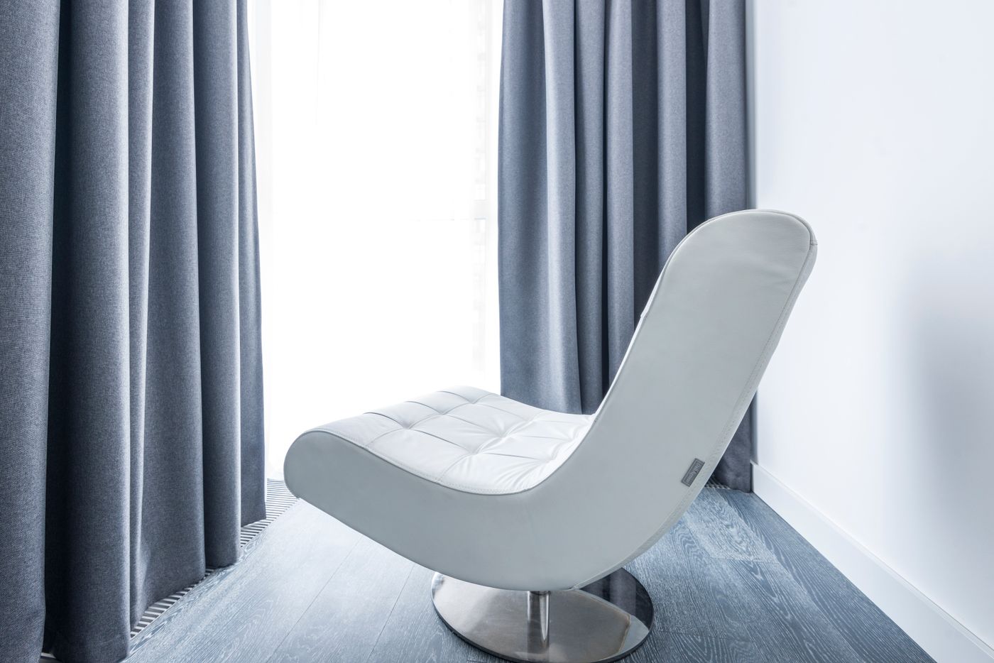 A white tufted lounge chair with a circular metal base beside gray curtains.