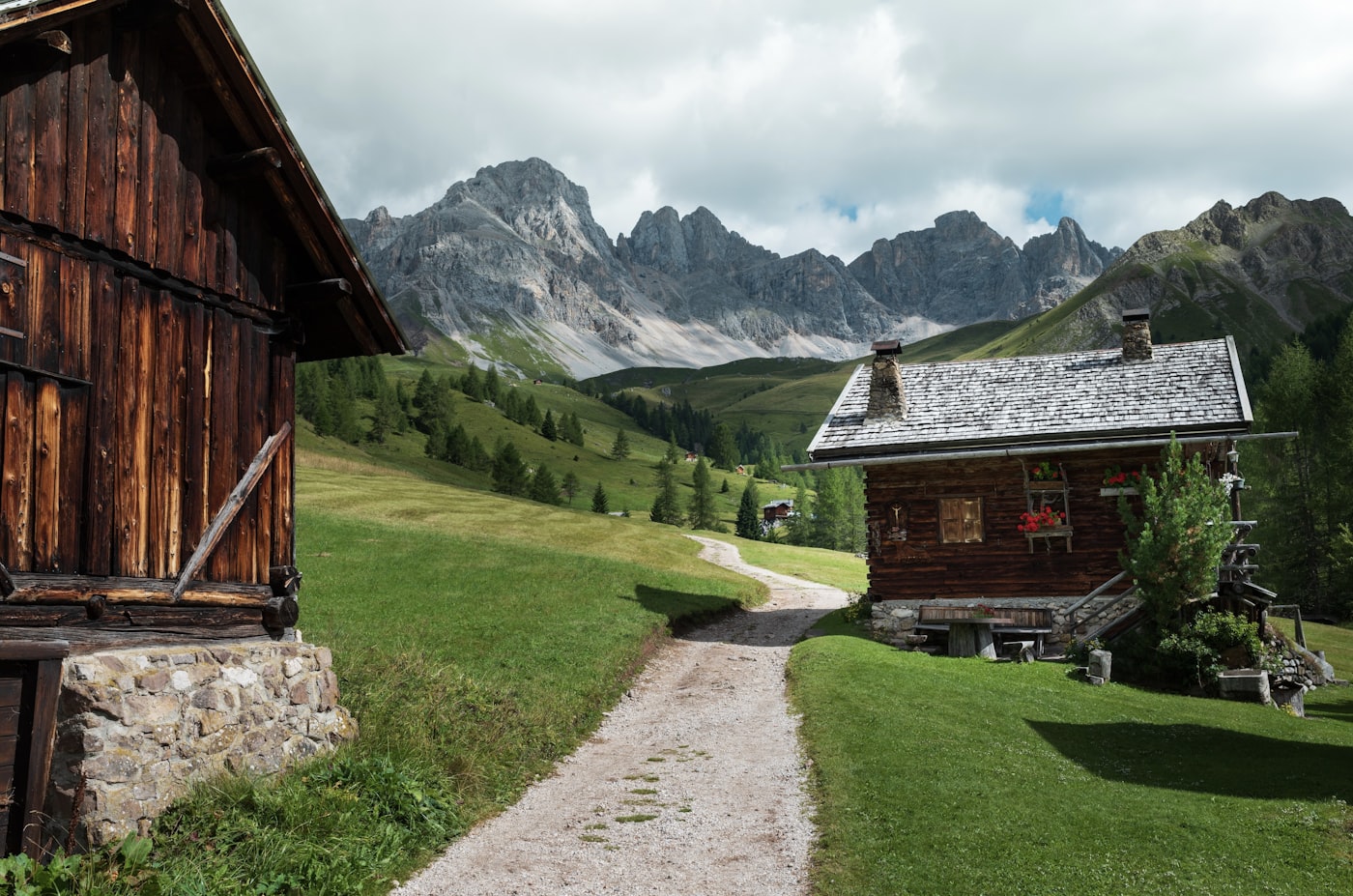 Wooden alpine cabins beside a meadow path beneath jagged mountain peaks